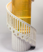 A close-up of a sculptural spiral staircase lamp or decor piece with a textured white spiral railing and a glowing warm gold vertical column behind it, set against a soft light gray background.
