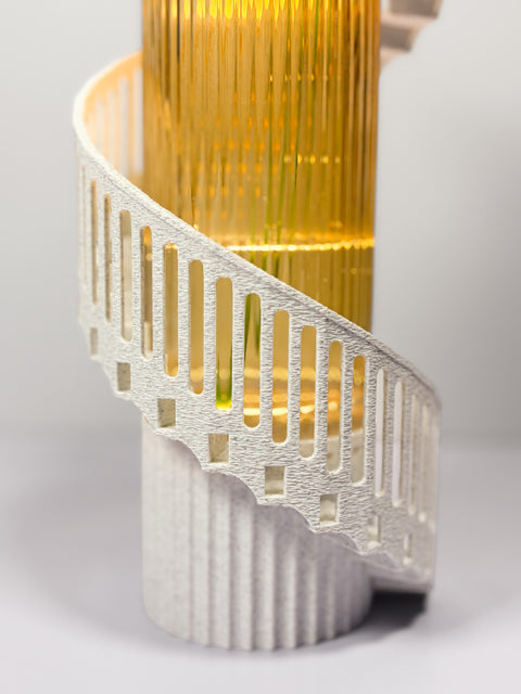 A close-up of a sculptural spiral staircase lamp or decor piece with a textured white spiral railing and a glowing warm gold vertical column behind it, set against a soft light gray background.