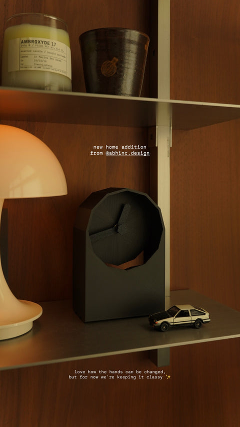 All black geometric clock sitting on a metallic shelf against a dark wood wall