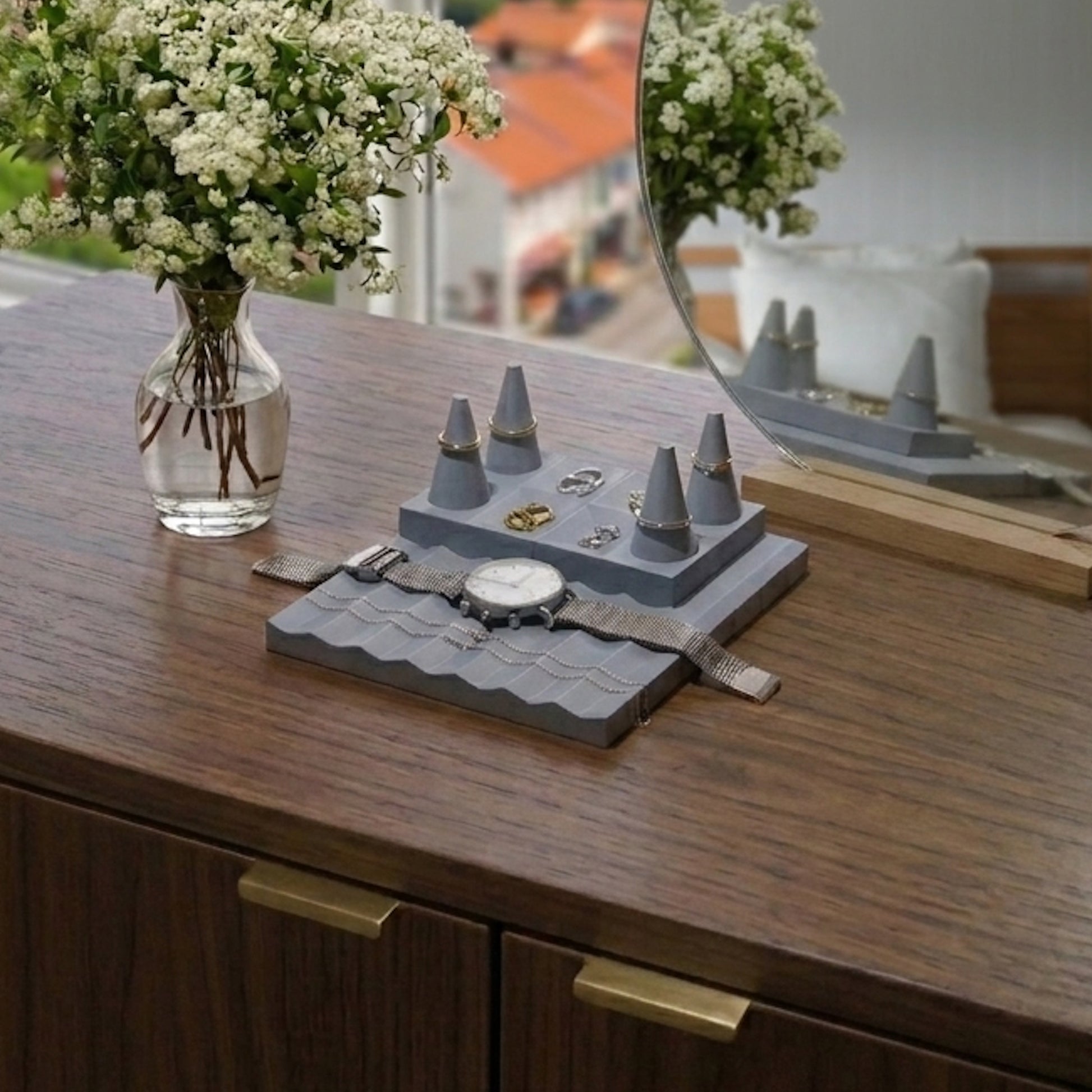 Gray jewelry display with watches and rings on a wooden surface, next to a vase of flowers and a mirror.
