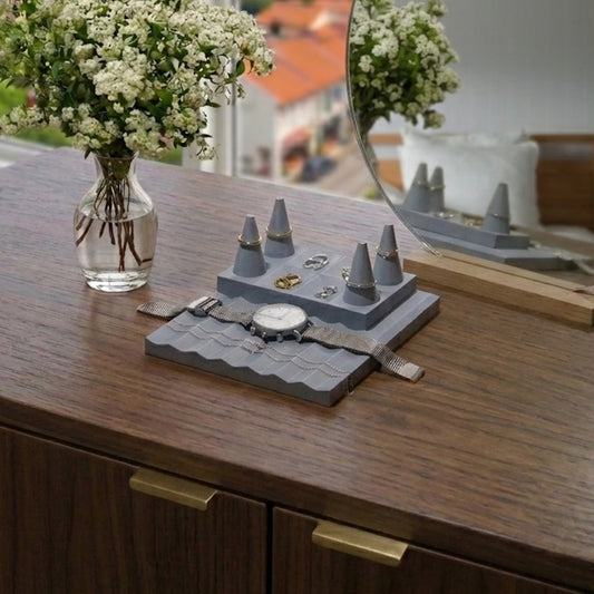 Gray jewelry display with watches and rings on a wooden surface, next to a vase of flowers and a mirror.